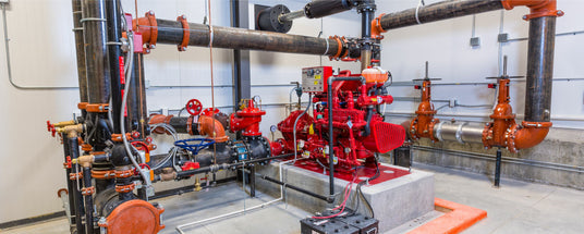 Fire sprinkler system equipment in an industrial setting, featuring red valves, black pipes, and a motorized pump on a concrete platform. The surrounding environment is a utility room with a white wall in the background.