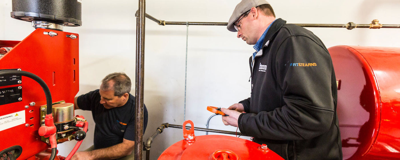 Two men working on industrial equipment. One is adjusting a red machine, while the other, wearing a cap and holding a device, observes. They are in a well-lit room with various pipes and machinery.