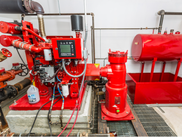 Fire control room with red industrial equipment, including a fire pump and control panel. Various pipes and valves are visible, along with a large red cylindrical tank in the background. A cleaning bottle is placed nearby.