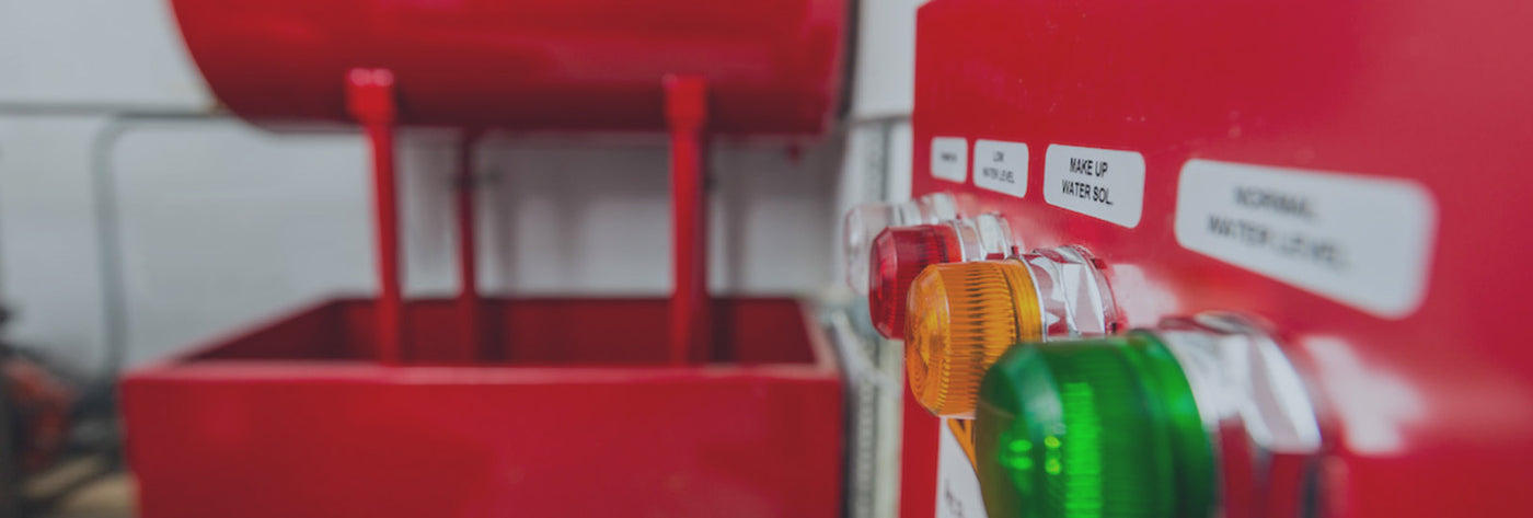 Close-up of a control panel with three illuminated lights: orange, red, and green. Labels are visible above each light. In the background, there is a large red container, slightly out of focus.