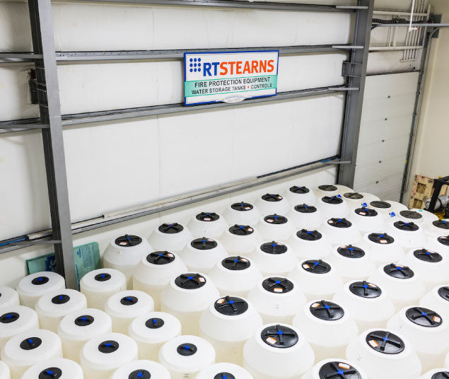 A warehouse with a large number of white cylindrical tanks stacked in rows. A sign above the tanks reads "RT Stearns Fire Protection Equipment Water Storage Tanks & Controls." The area is well-lit and organized.