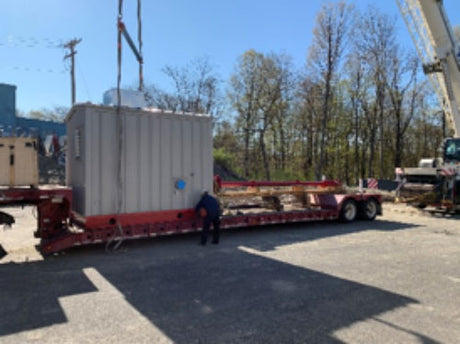 A large flatbed trailer is parked outdoors, holding a gray shipping container that functions as a fully operational Patterson Prepackaged System fire pump house. A person stands beside it, with trees and a crane visible in the background under a clear sky.